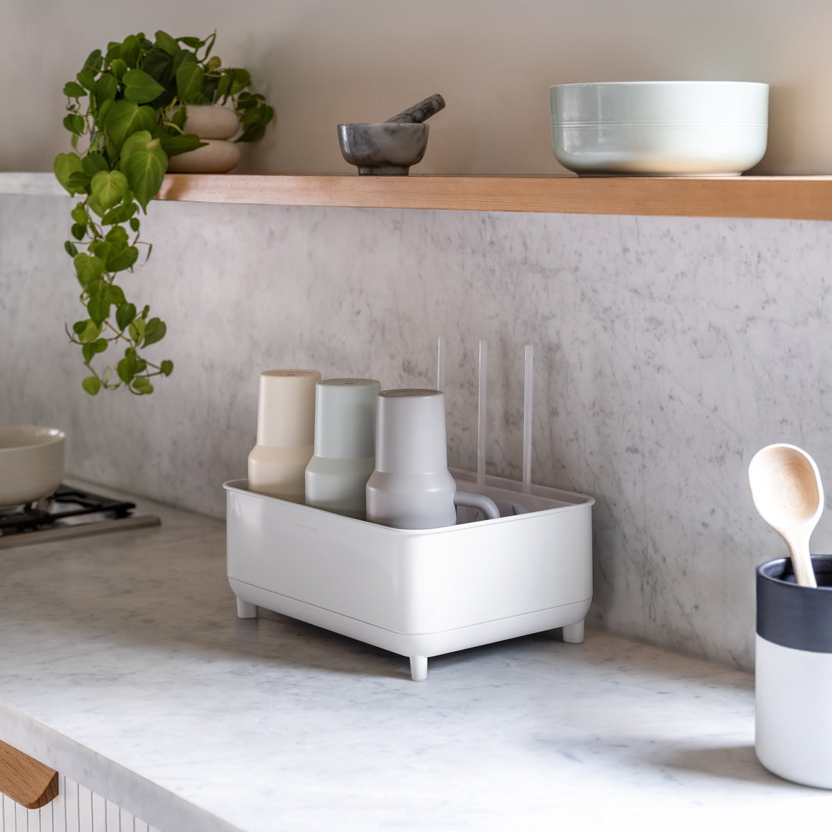 Simplehuman White Bottle Dock on marble kitchen countertop holding bottles and mugs beside a plant and utensil holder.