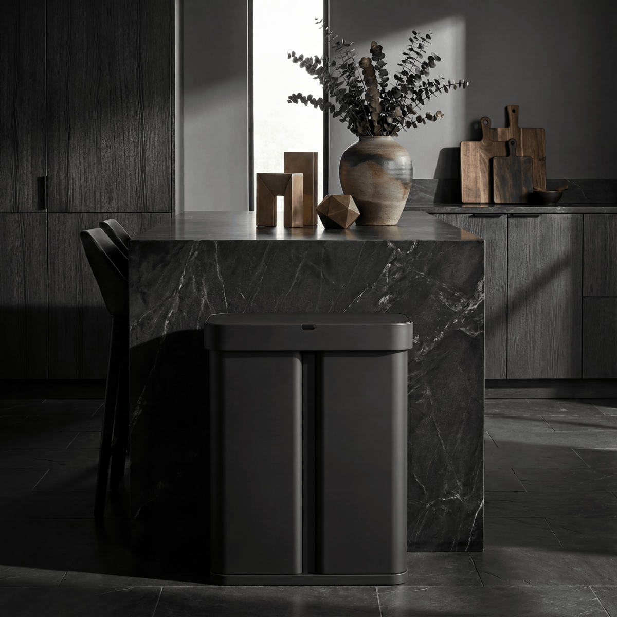 Simplehuman's 58L Sensor Can in Matte Black in a darker background with black countertop and cabinets featuring warm, wooden trinkets on the countertop and cutting boards in the background.